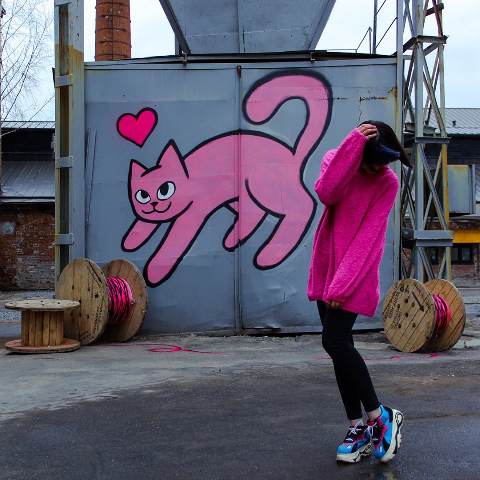 Woman in pink sweater dancing in front of a large pink spraypainted cat on a wall